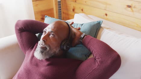 happy senior african american man in log cabin, laying on sofa and using headphones, slow motion