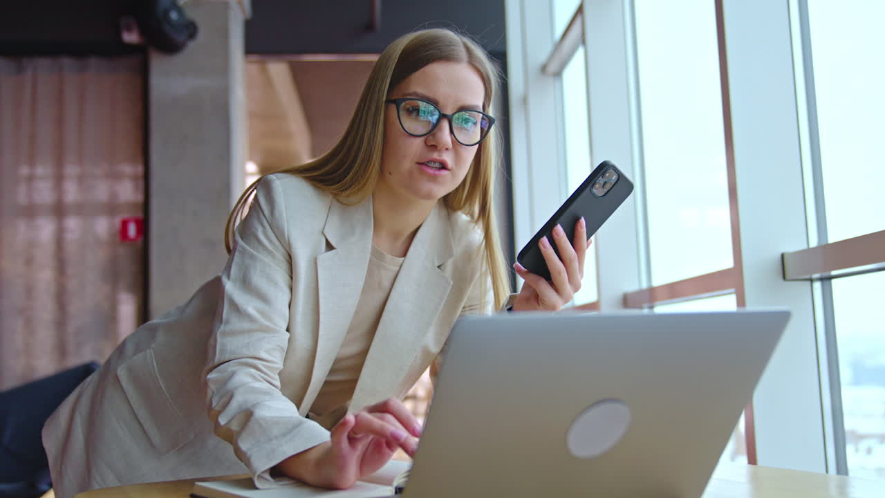Blonde young woman speaking on the phone leaning on the table. Female employee talks on the phone and works on laptop. Office at backdrop.
