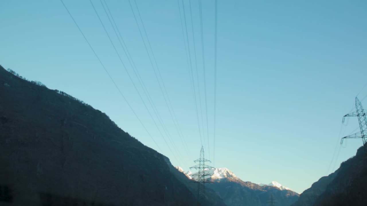 Car POV of power lines stretching over mountains with snowy peaks in the distance under a clear sky