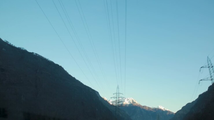 Car POV of power lines stretching over mountains with snowy peaks in the distance under a clear sky