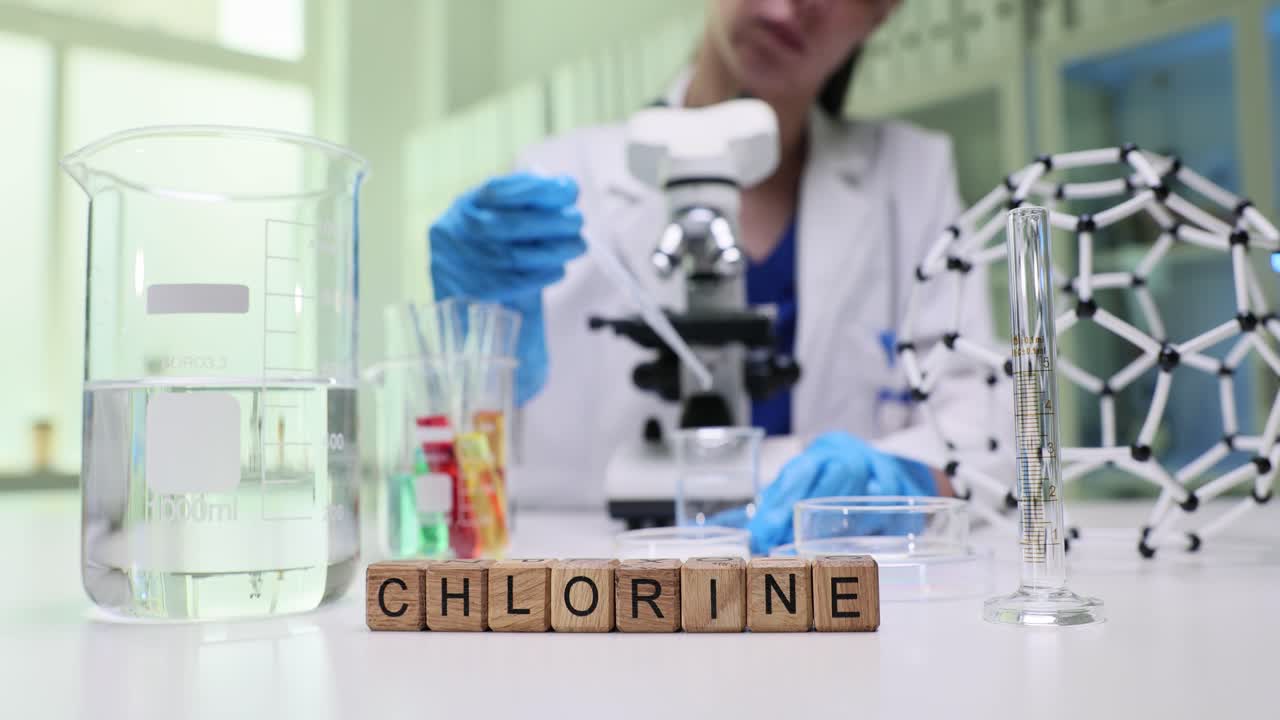 A person in a lab coat conducting a chemistry experiment in a laboratory, with the word 'CHLORINE' spelled out in wooden blocks