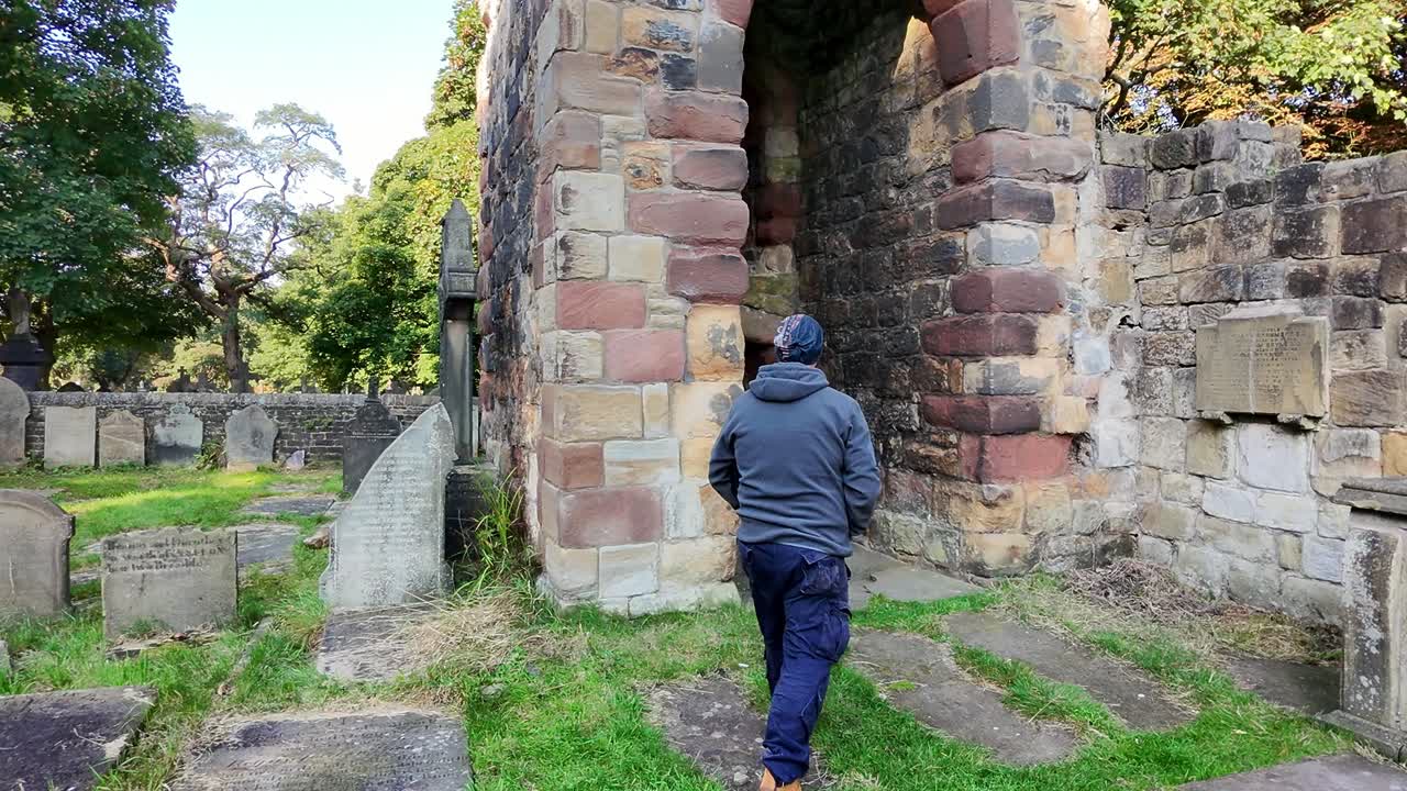 Following male wearing hat and casual clothing walking alone in peaceful sunlit cemetery abbey ruins