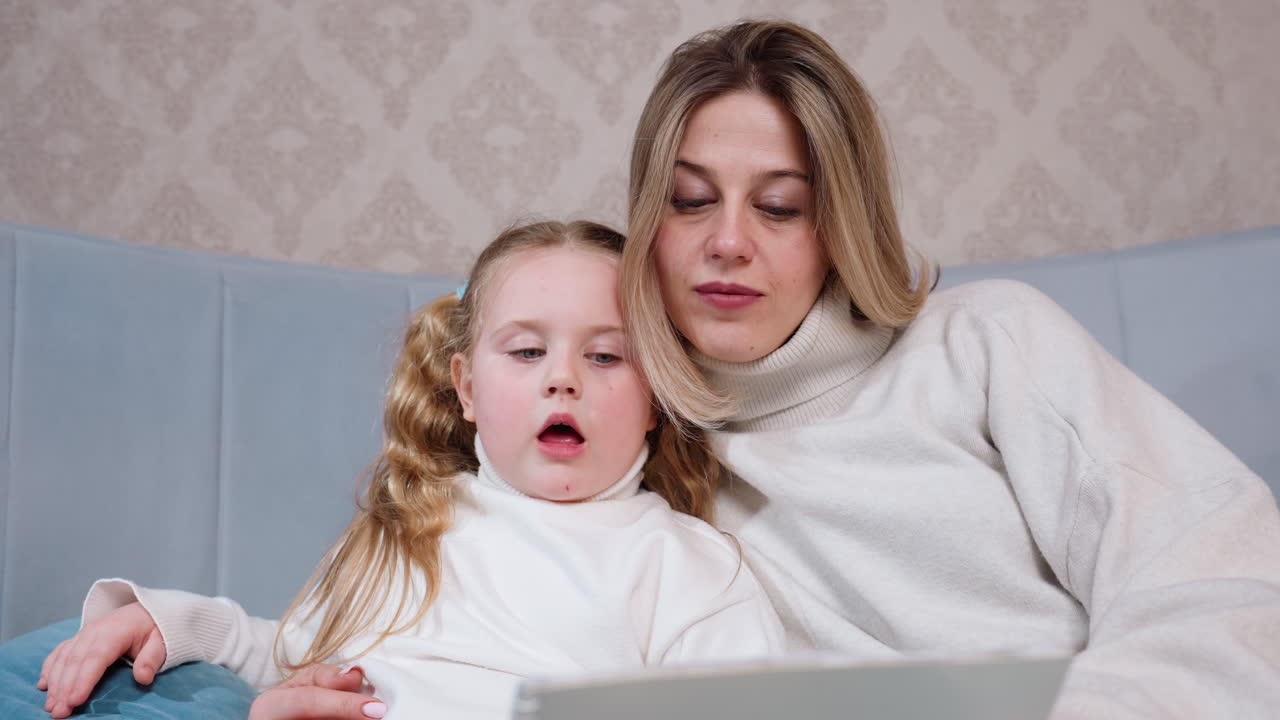 Smiling young girl with long blonde hair and woman in cozy sweaters sitting close on couch looking at sketchbook together, enjoying creative drawing activity and spending warm bonding time