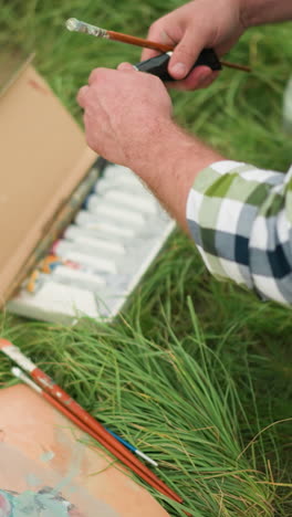 un dibujante en una camisa a cuadros se inclina en un campo de hierba, abriendo cuidadosamente un tubo de pintura de una caja de pinturas. una paleta con varios colores también está en la hierba