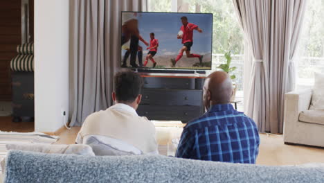 Vista-Trasera-De-Un-Padre-Afroamericano-Y-Un-Hijo-Adulto-Hablando-Y-Viendo-Rugby-En-La-Televisión,-Cámara-Lenta