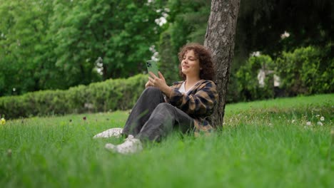 chica feliz con el cabello rizado en una camisa a cuadros escribiendo en su teléfono inteligente verde y comunicándose en redes sociales en el parque