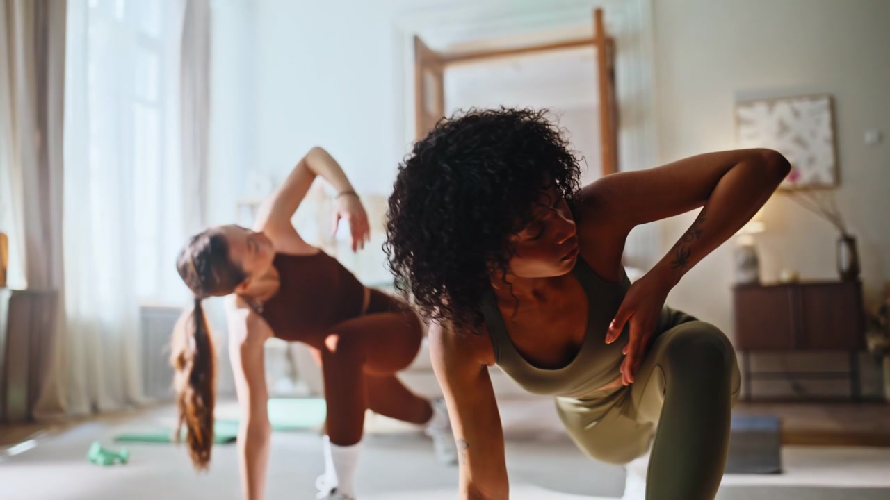 Multiracial friends home workout. Two women stretching side lunges living room.