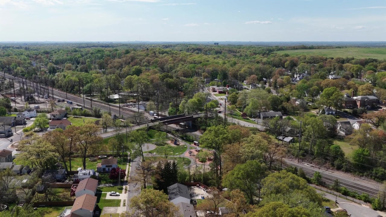 Cittadina suburbana americana con tetti di case e una ferrovia che attraversa un paesaggio primaverile verde sotto un cielo azzurro limpido, con un'atmosfera di quartiere tranquillo e un ambiente panoramico. Vista aerea con drone.