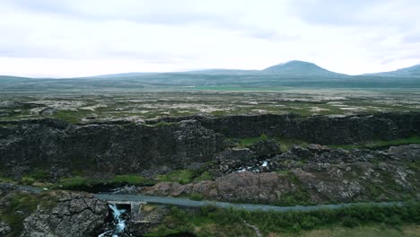 Iceland,-road-under-rocks-and-rock-cliff-with-small-waterfall,-aerial-video