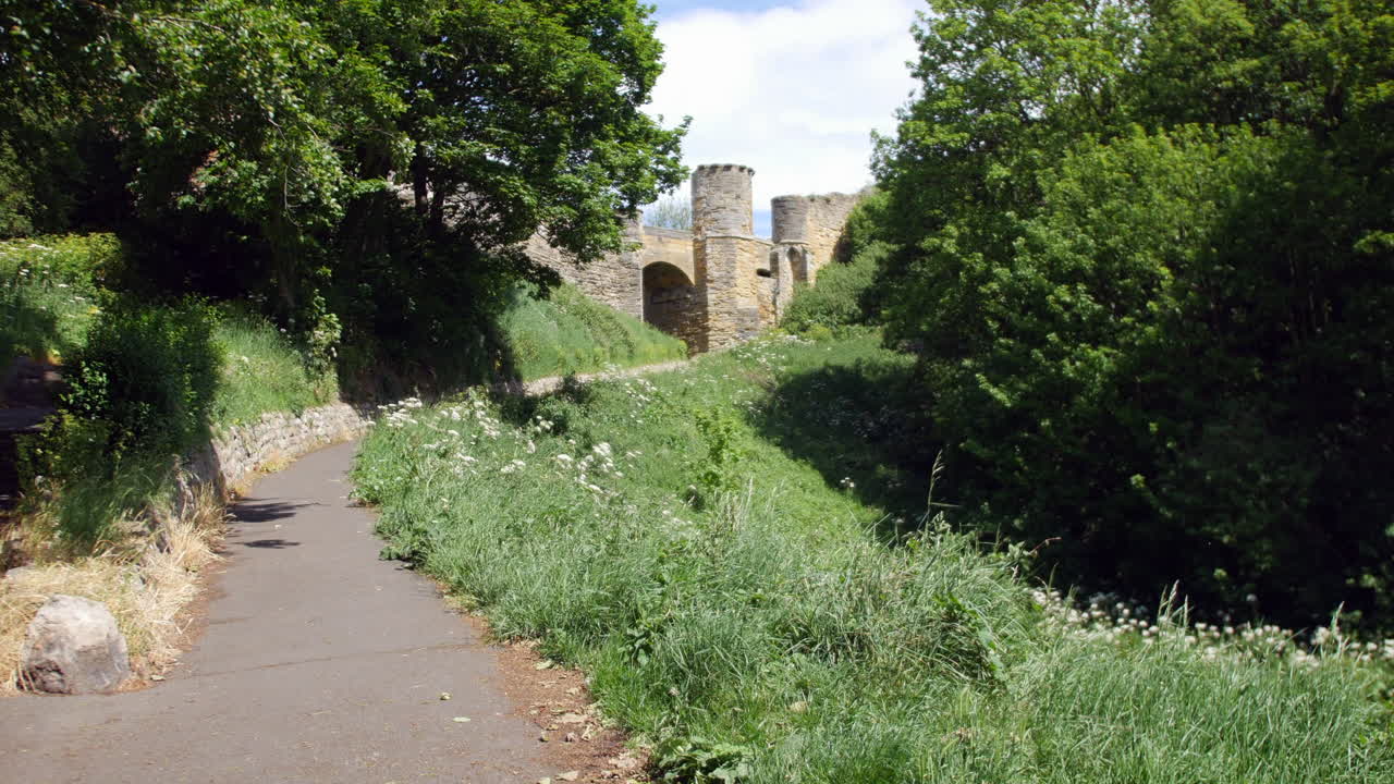 A winding path lined with grass and flowers leads toward castle ruins in Scarborough, North Yorkshire, England, with ancient stone towers rising above