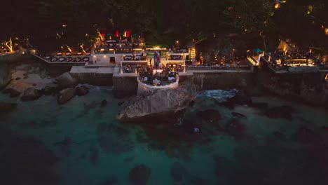 a unique bali bar perched on a rocky cliff welcomes diners at sunset time where people relax over meals listening to the ocean and soaking in the calm atmosphere of the evening