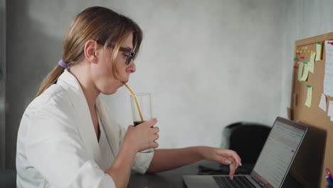 remote worker wearing glasses drinking dark beverage with straw while typing on laptop with left hand seated in modern home office setting, corkboard full of colorful sticky notes on board