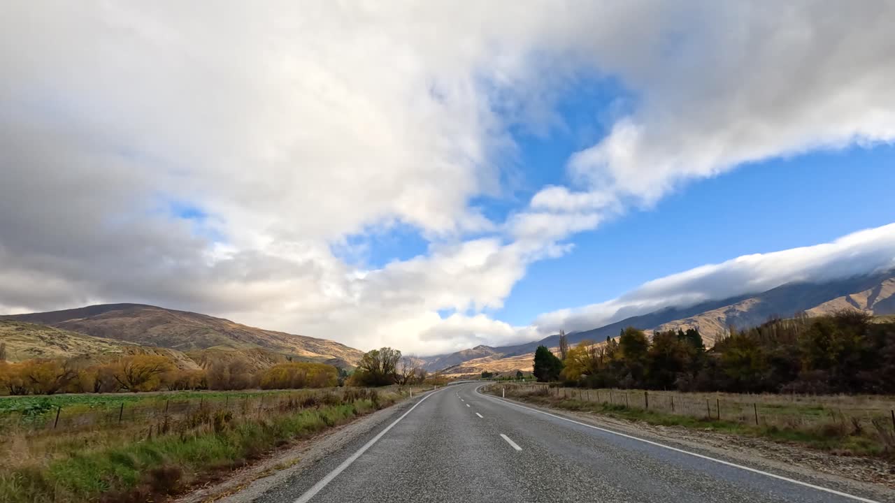 A serene drive through Wanaka's countryside, showcasing vibrant autumn foliage, expansive skies, and winding roads under dynamic cloud cover