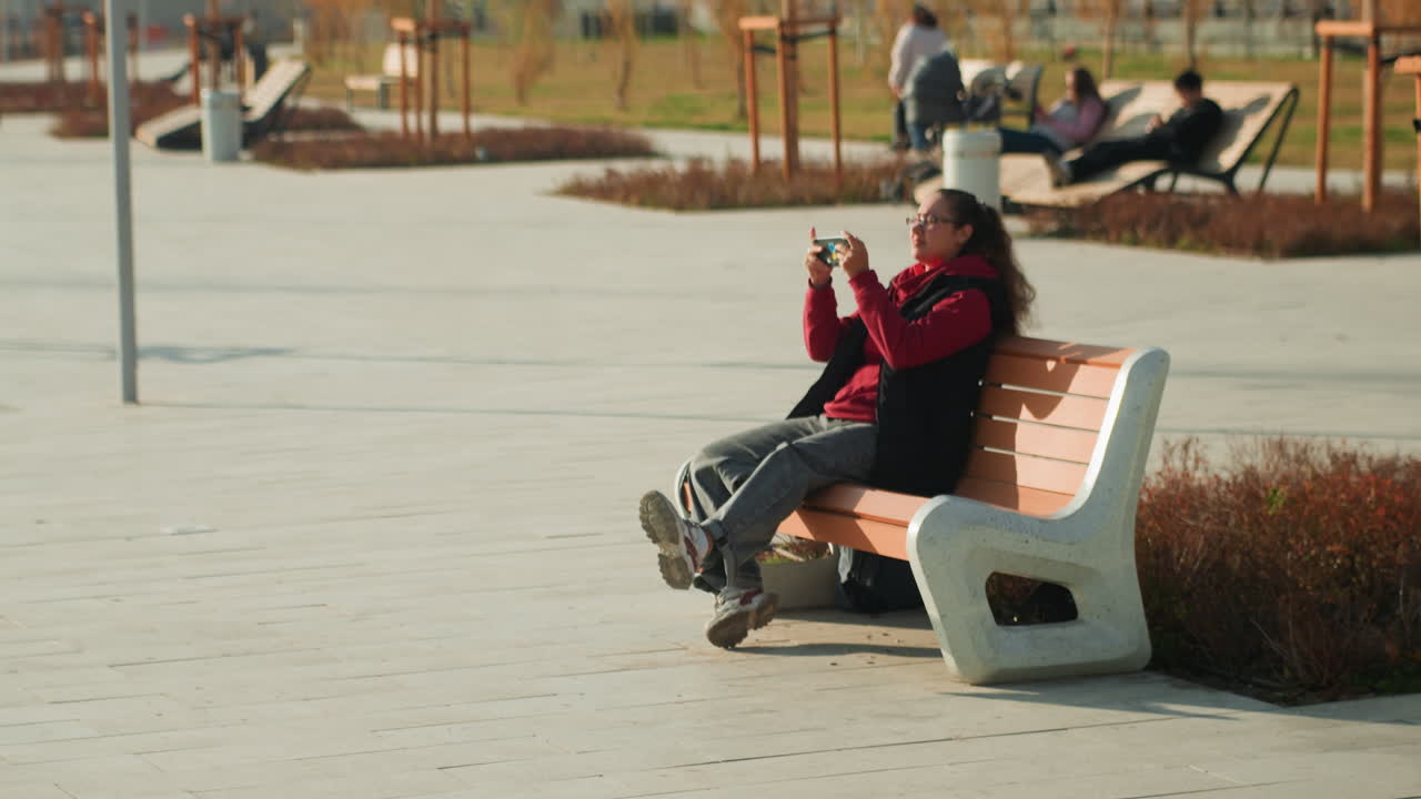 Donna afroamericana seduta su una panchina in un parco urbano, con una giacca rossa logora, la testa tra le mani, il telefono riposto. Freddo autunnale, piazza di cemento, passanti, edifici distanti, luce soffusa, umore introspettivo e ansia.