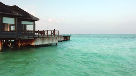 Aerial,-drone-shot,-low-over-turquoise-ocean,-towards-a-girl,-walking-on-the-balcony,-at-a-villa-on-the-sea,-on-a-sunny-day,-on-the-Conrad-Rangali-island,-in-Maldives