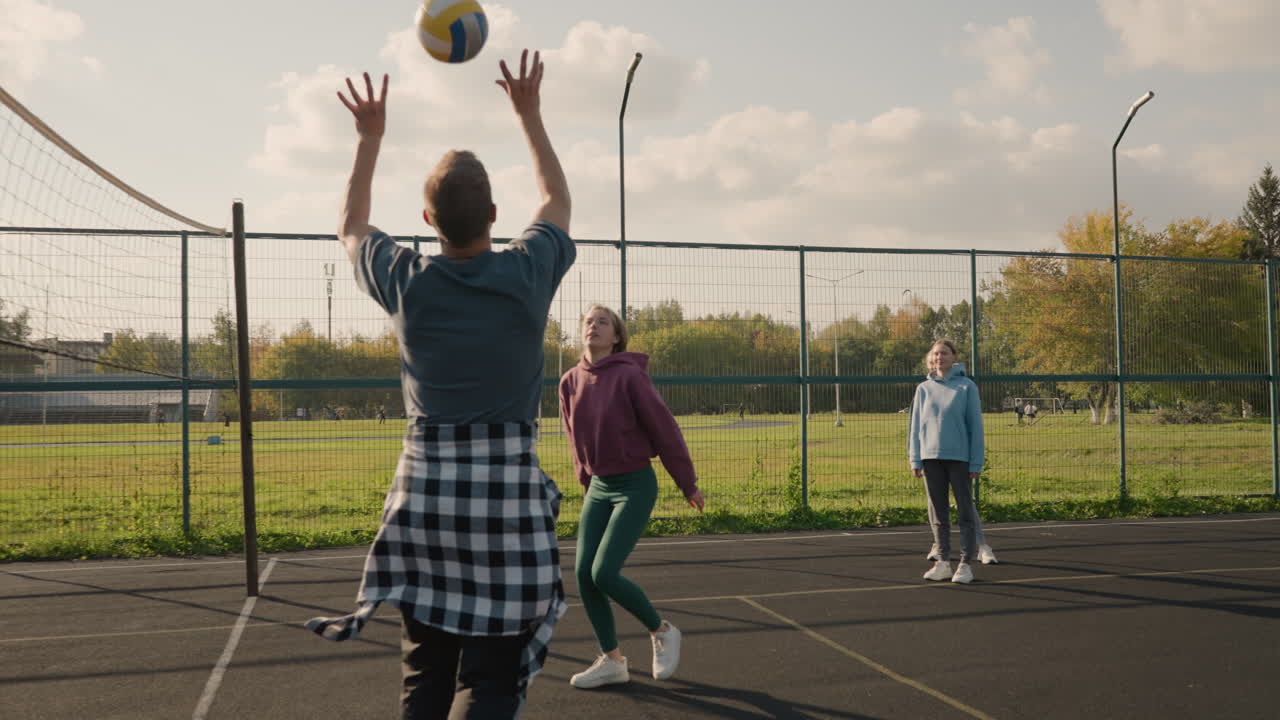 두 명의 다른 팀 동료가 지켜보는 동안 코치와 함께 배구를 하는 여자, 야외 스포츠 코트 위의 축구 경기장과 은 하늘을 볼 수 있습니다.