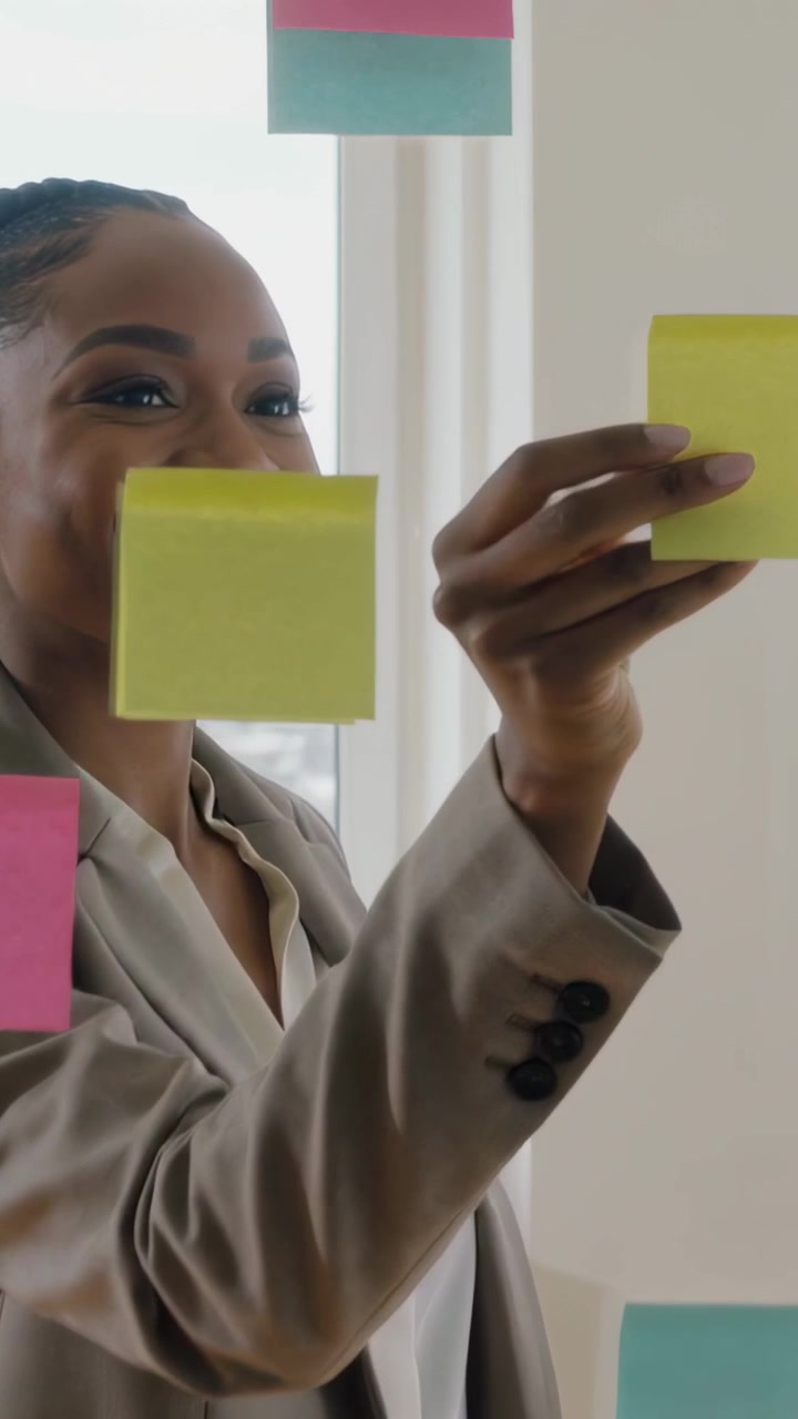 Video verticale: Afferrando un blocco di post-it, una donna in blazer posiziona un post-it giallo sul vetro dell'ufficio, pianificando.