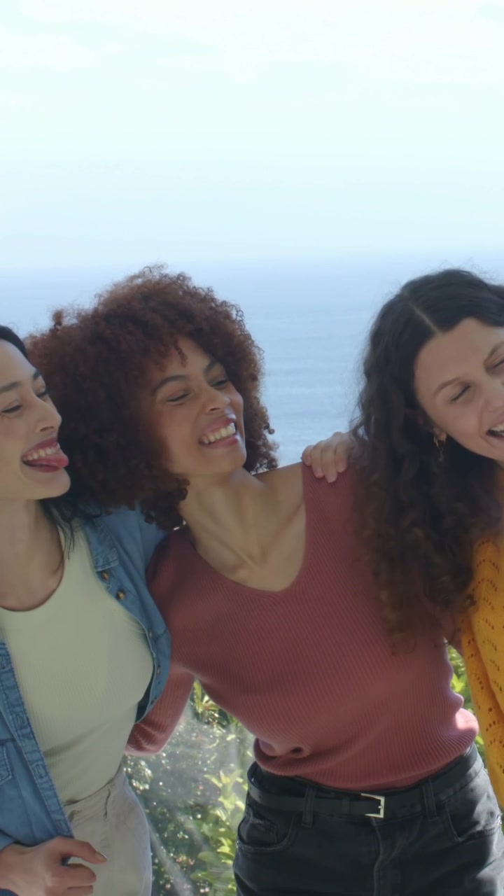 Video vertical: Tres amigos apoyados posando de forma juguetona para la cámara en un balcón junto al mar, con suéteres