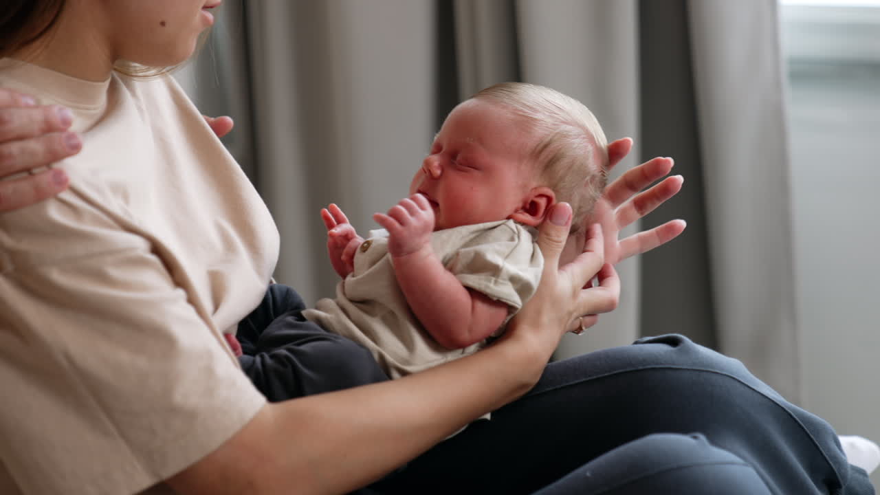 Adorable tiny baby boy in beige t-shirt and pants. Infant starts to wake up and mom changes his position calming him down.