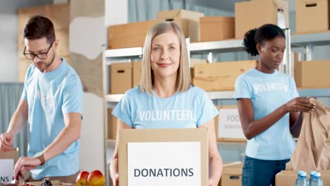 caucasian woman volunteer holding donation boxes and looking at camera in charity warehouse while her coworkers working packing boxes