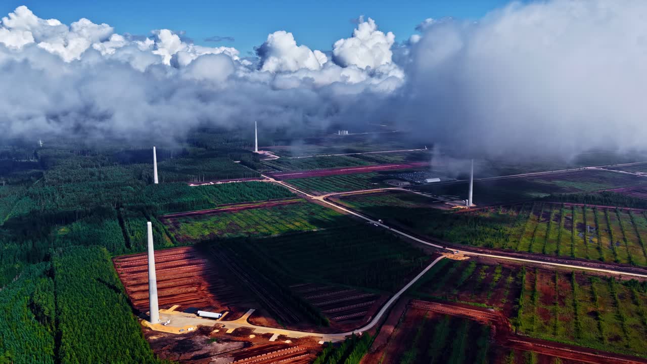 Aerial overview of wind farm construction site with tall turbine towers amid green fields forests roads and drifting clouds over Latvian countryside