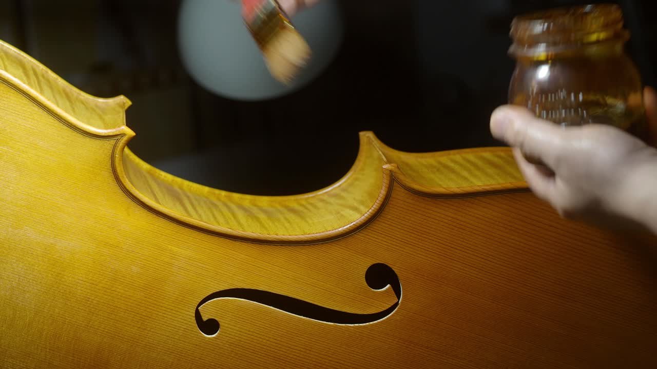 Close-up of a luthier applying varnish to cello ribs and edges on center bout near f-hole, highlighting the wood grain and f-hole detail in warm light