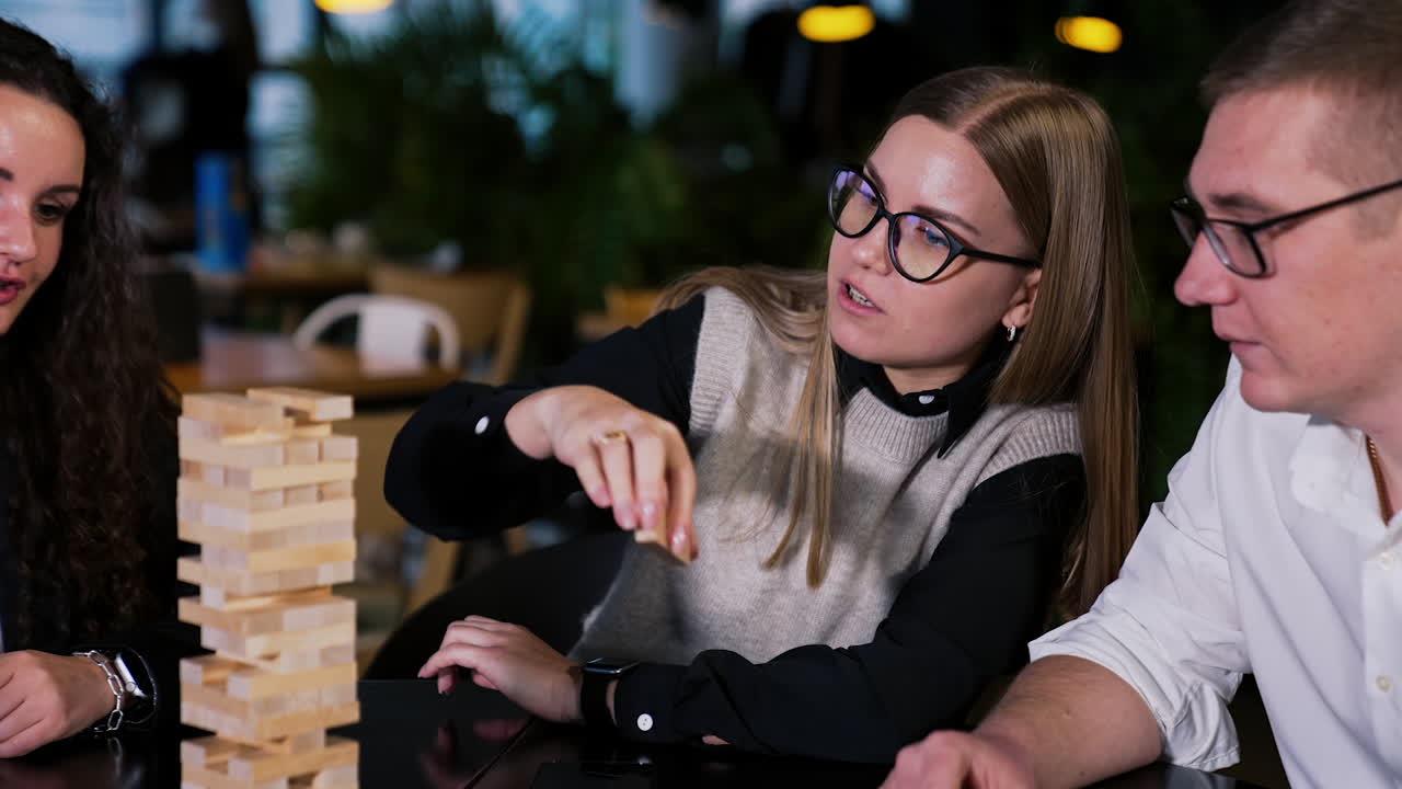 Friends playing jenga game and talking. Long-haired blonde girl pulls the wooden brick and puts it on top of a pile.