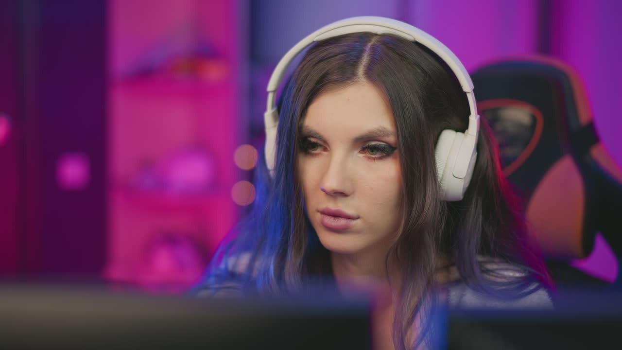 young female programmer wearing white headphones works in front of computer screen with serious expression, seated in gaming chair surrounded by blurred colorful equipment in modern studio setting