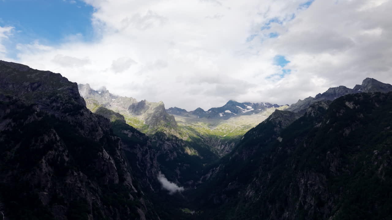 Bidikan drone udara perlahan bergerak maju dengan gerakan menggeser di atas lembah pegunungan di Pegunungan Alpen Swiss, menampilkan lereng yang teduh dan terkena sinar matahari, awan, dan langit yang dramatis