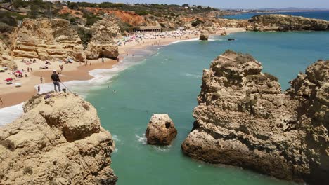 Fischer-Steht-Auf-Felsen-Im-Vordergrund,-Strand-Von-Albufeira-Im-Hintergrund,-Portugal,-Hoher-Winkel