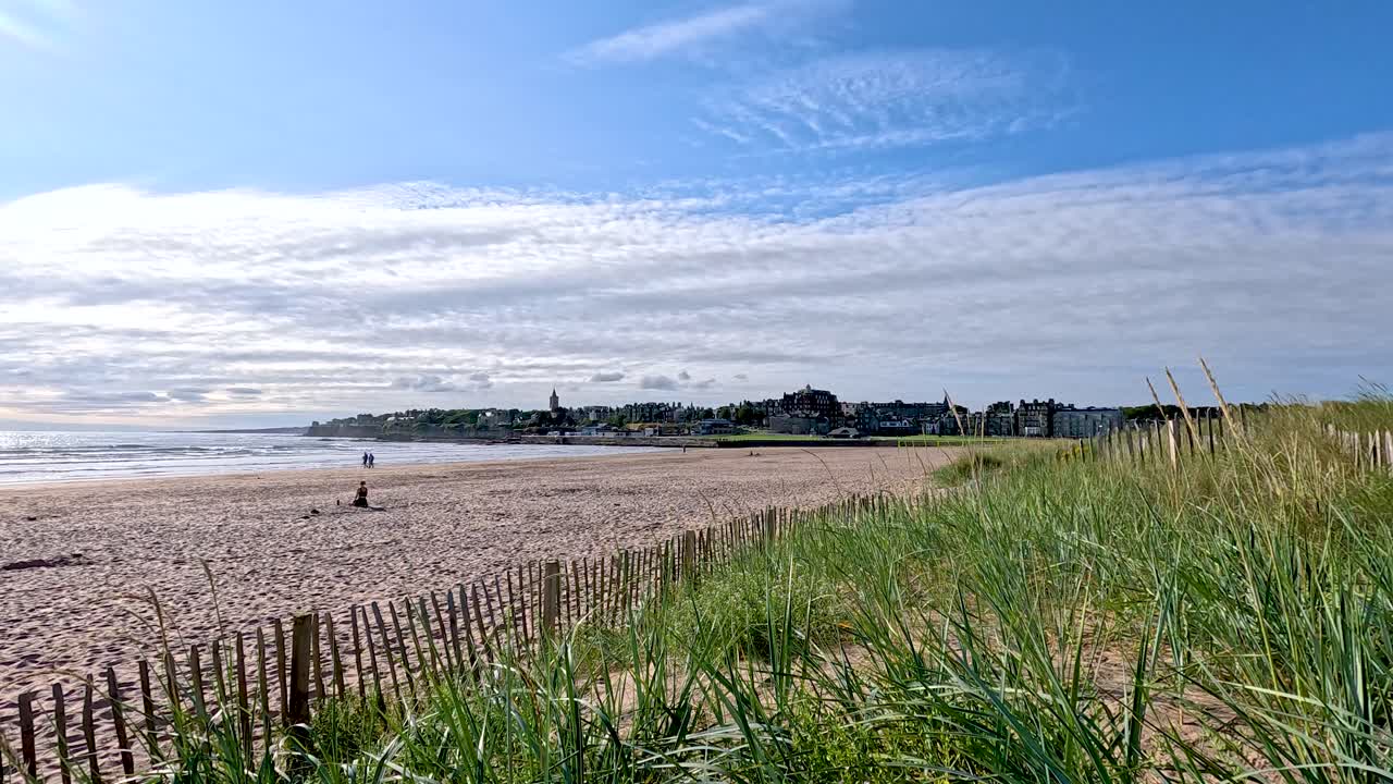 Camera pans from grassy sand dunes, revealing beach, wooden fence, blue sky, and distant town