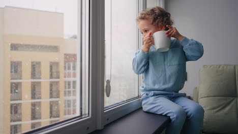 cute slavic little boy with cup is looking out window sitting on sill in winter day modern apartment in high building