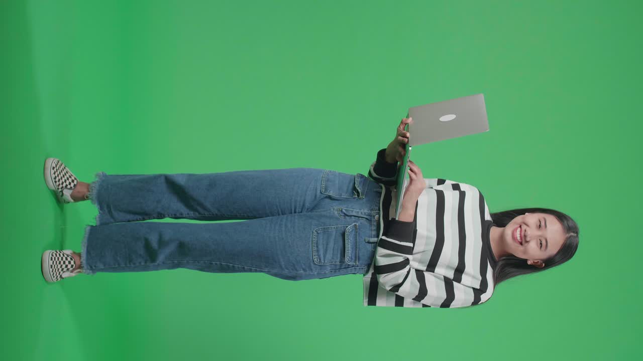 cuerpo lleno de una mujer asiática sonriente mirando a la cámara mientras escribe en la computadora en el estudio de pantalla verde