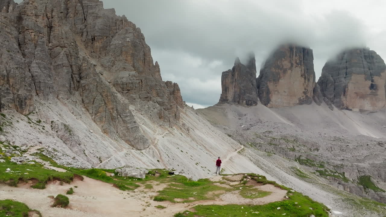 Drone circles person standing at edge looking out to Tre Cime di Lavaredo mountain with distinct rugged spires in Dolomites