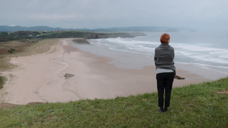 woman stands on a grassy clifftop overlooking a vast sandy beach and ocean