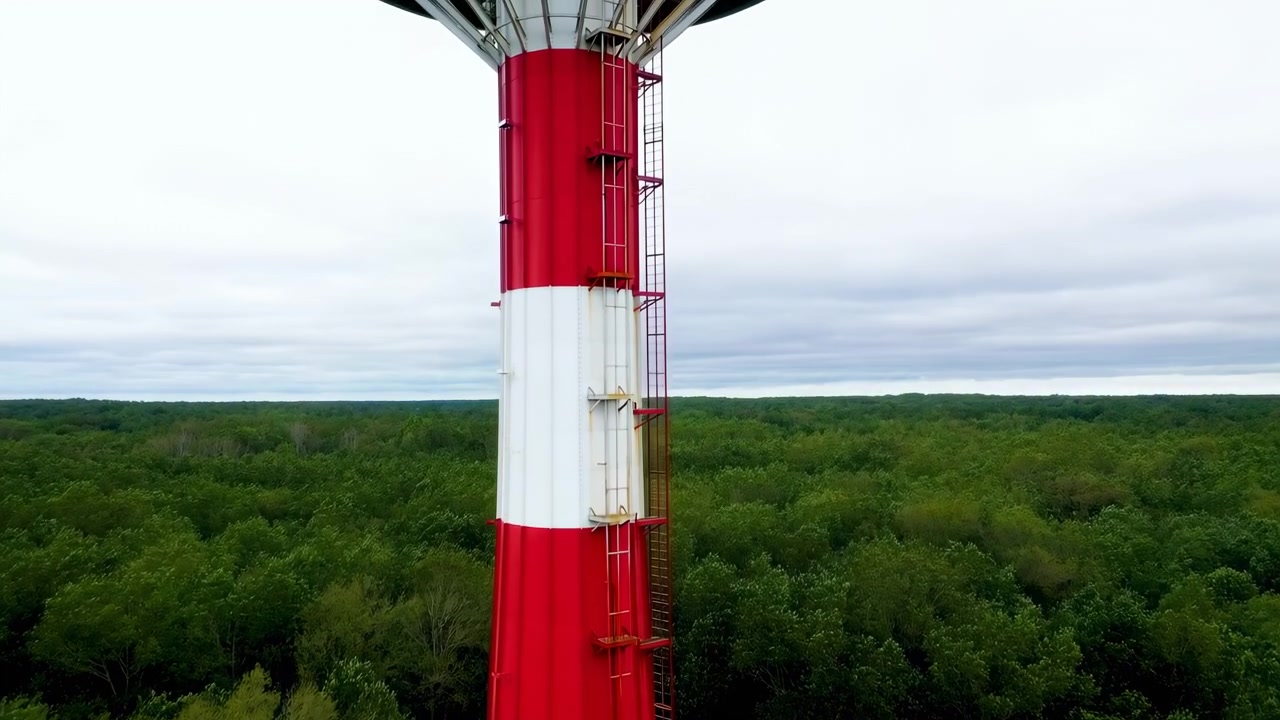 Menara merah dan putih besar menjulang di atas hutan hijau lebat, dengan tangga vertikal, pagar pembatas, dan detail industri yang lapuk menonjol di langit berawan dalam lanskap pedesaan terbuka