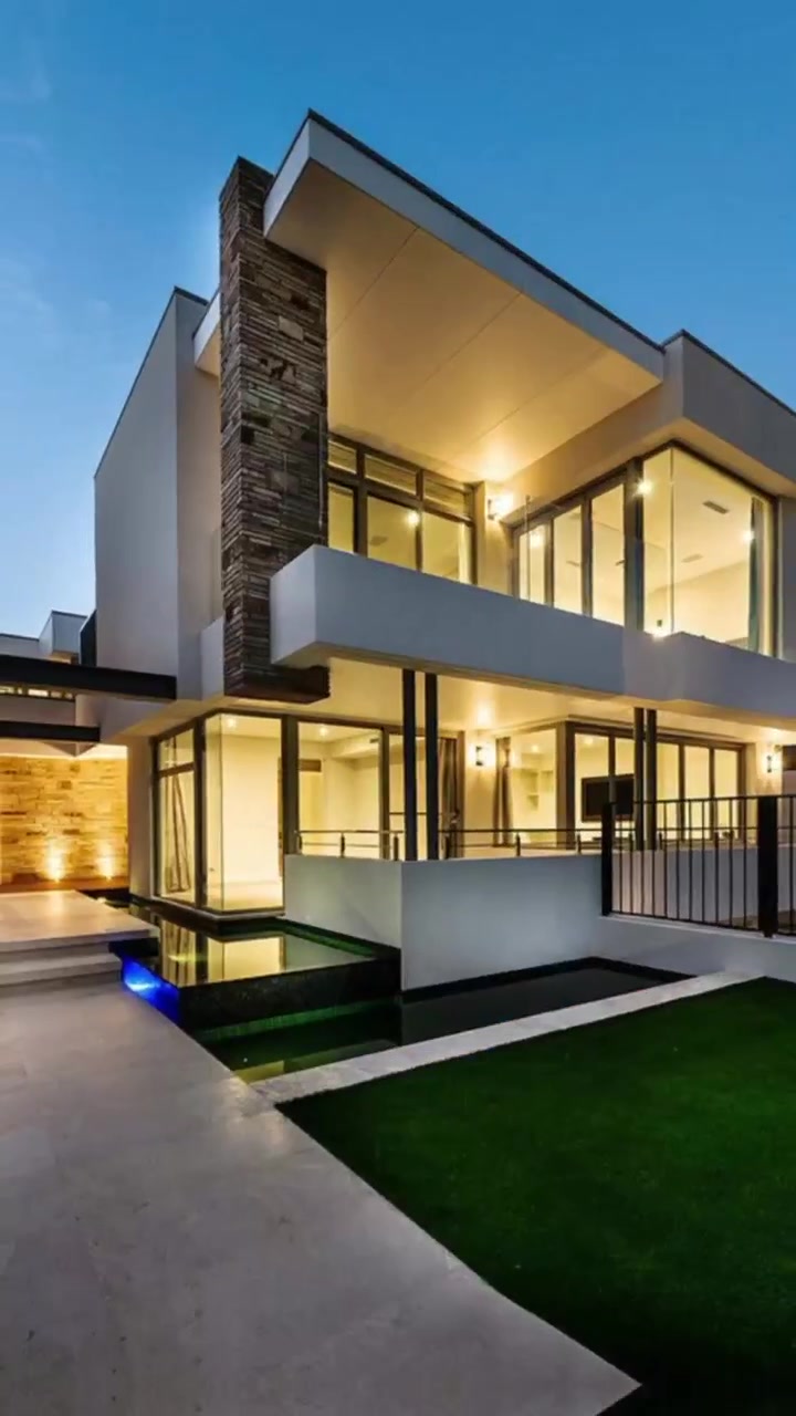 Exterior de casa de lujo moderna con ventanas iluminadas y fuente de agua al anochecer