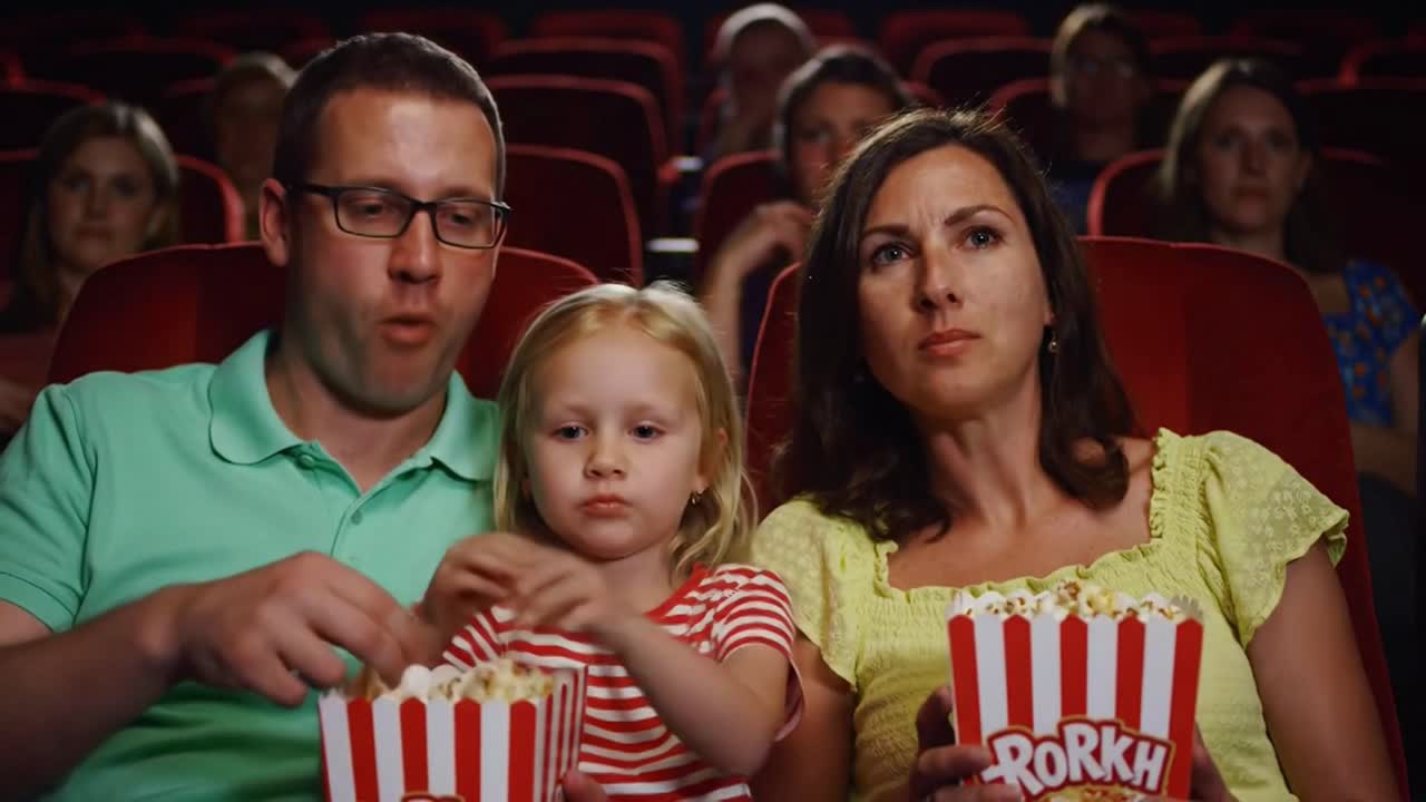 A family, mother, father, and young daughter, eat popcorn while intently watching a movie in cinema.