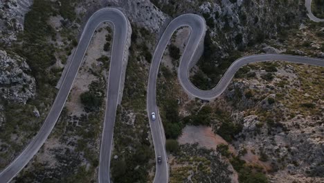 aerial drone top down shot over cars passing along winding sa calobra road over rocky mountain range in mallorca, spain during evening time