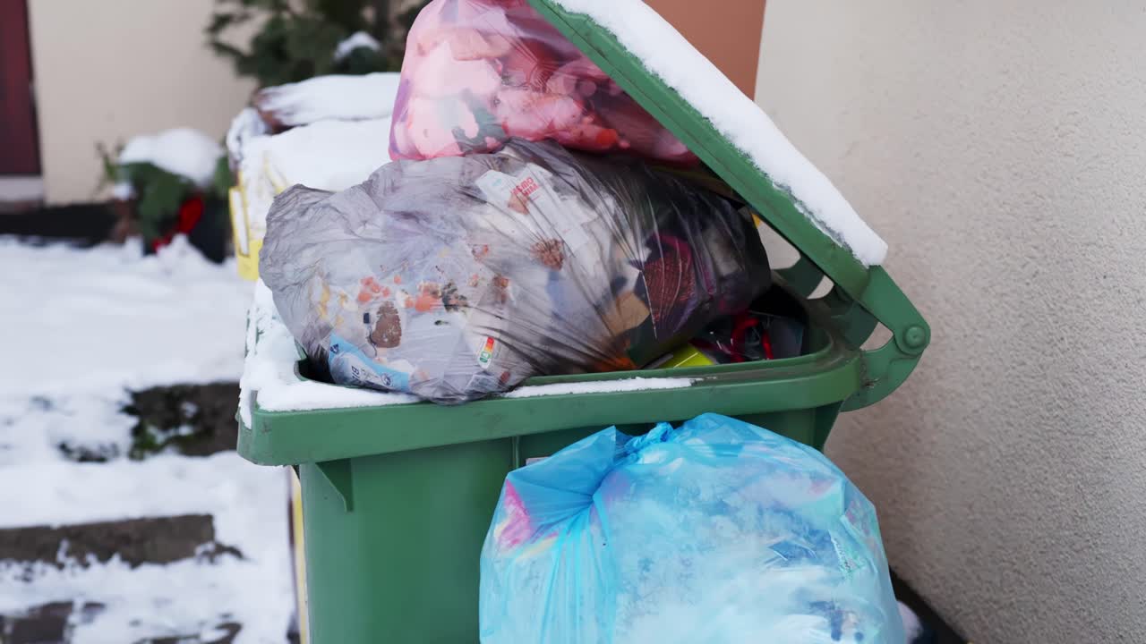 Cestino della spazzatura stracolmo di sacchi di immondizia sotto la neve accanto a una casa, che mostra problemi di igiene invernale, rifiuti e inquinamento urbano in una zona residenziale
