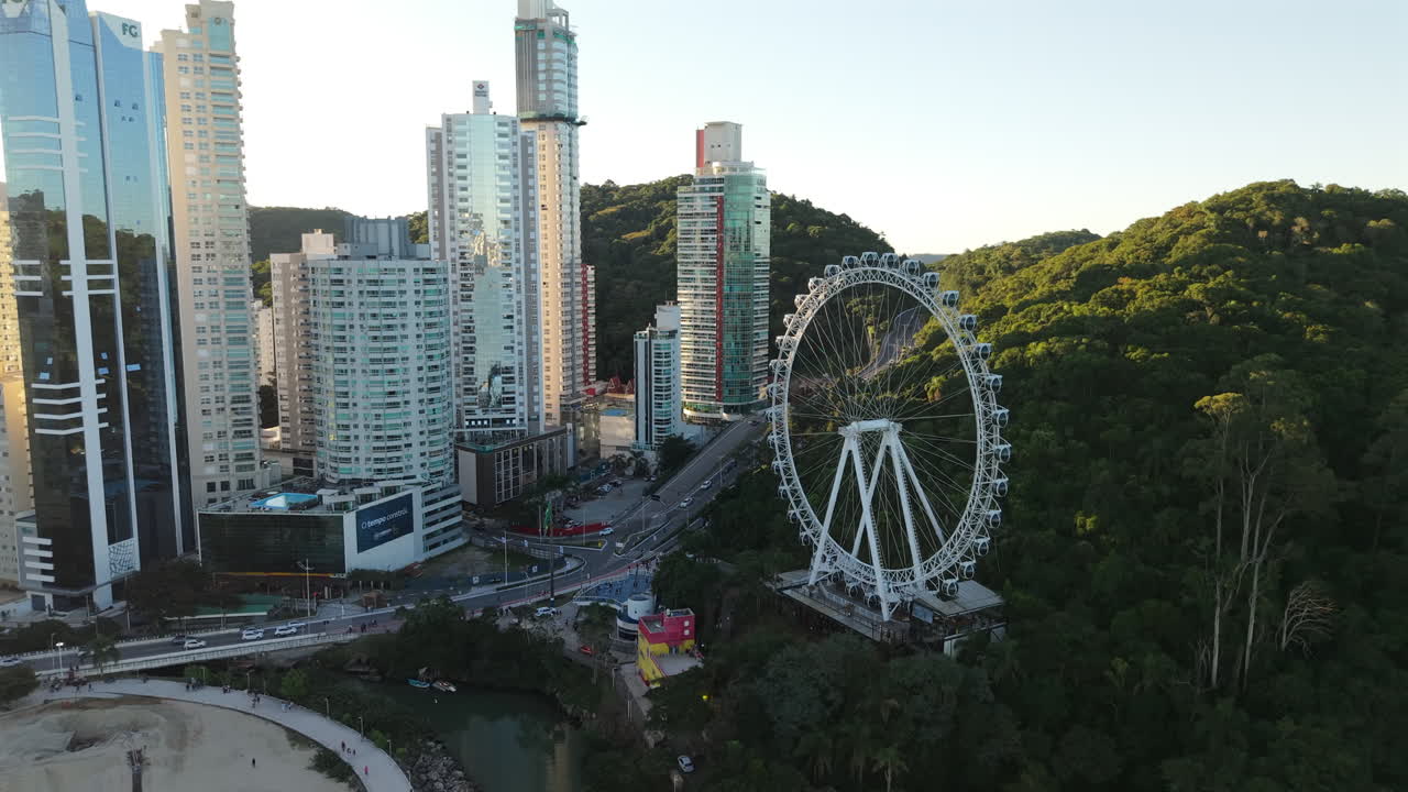 Drone shot of Balneario Camboriú’s white ferris wheel near office buildings, green hill, and ocean. Afternoon light, Brazil