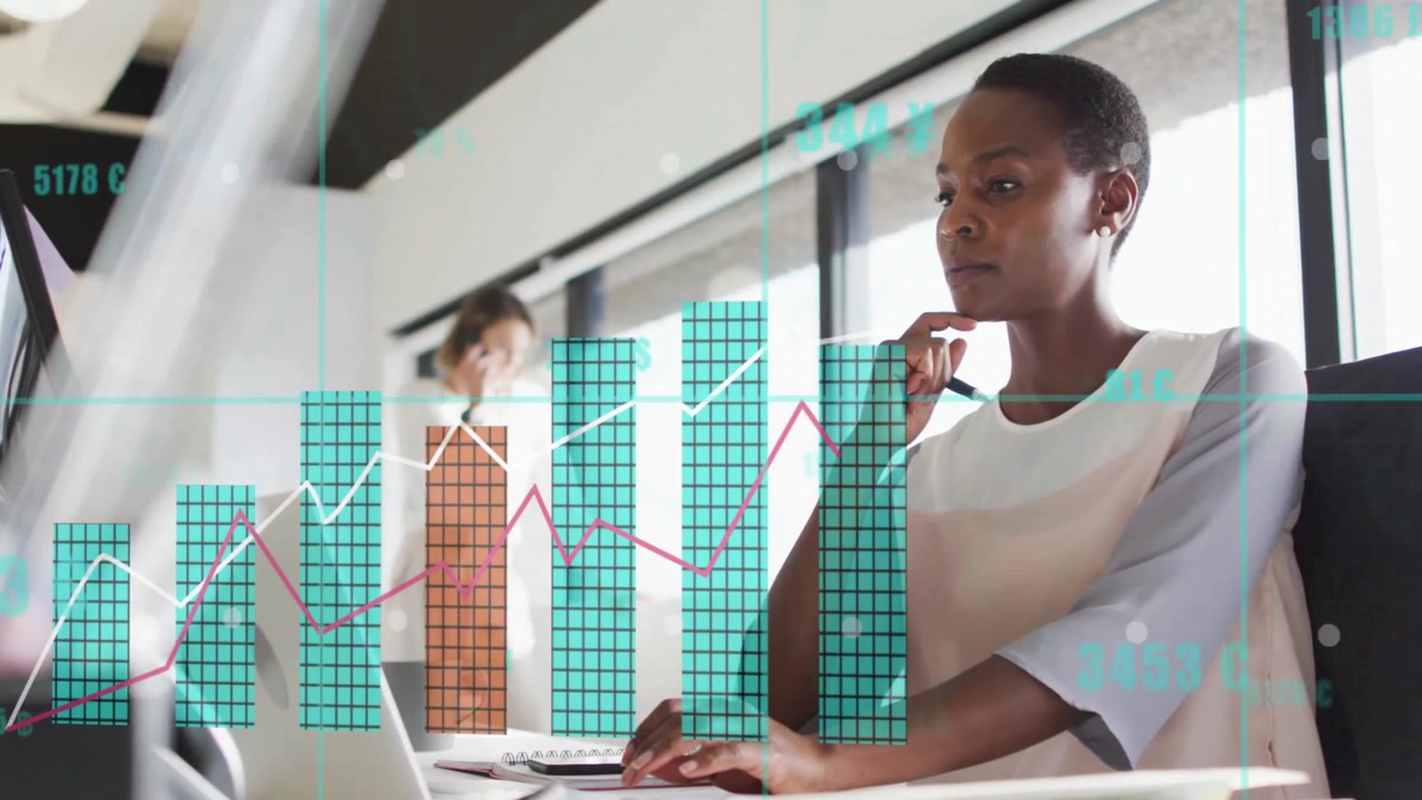 Woman reviewing overlays on laptop in office while coworker calling on phone showing finance charts