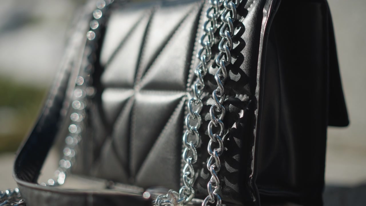 Primo piano di una borsa in pelle nera con tracolla a catena argentata appoggiata su una superficie di cemento. L'illuminazione soffusa evidenzia la texture trapuntata e i dettagli metallici lucidi creando un effetto bokeh sullo sfondo