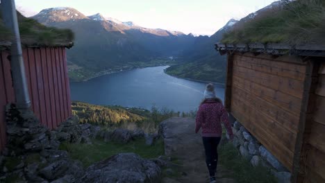 niña caminando entre dos cabañas pequeñas y antiguas, hacia el punto de vista, observando un hermoso paisaje de montañas y un gran lago en loen, noruega