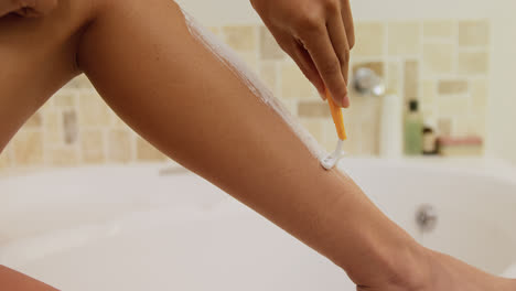 Young-woman-sitting-on-bathtub-waxing-her-leg-with-razor-in-bathroom-4K-4k