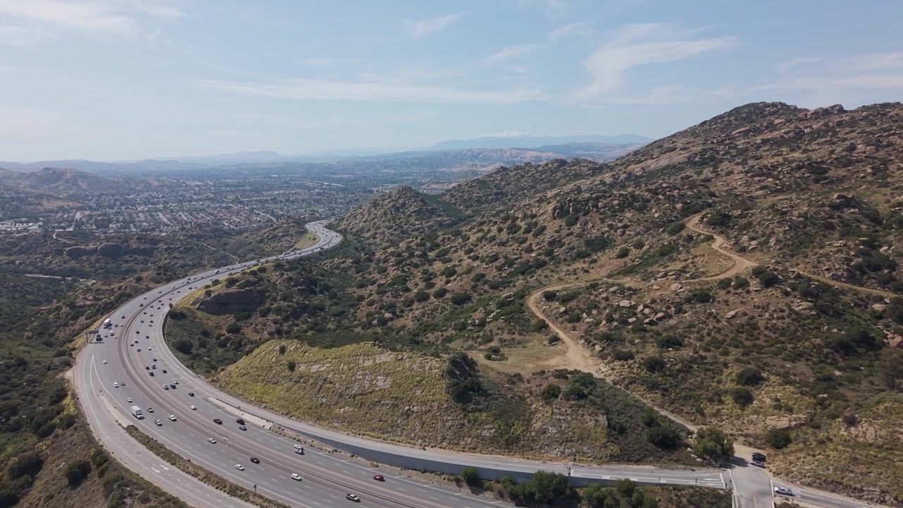 Autostrada a più corsie con traffico costante che attraversa colline aride nella Simi Valley, California, sotto un cielo azzurro limpido, con sentieri sterrati tortuosi, terreno accidentato e paesaggio suburbano. Ripresa panoramica aerea.