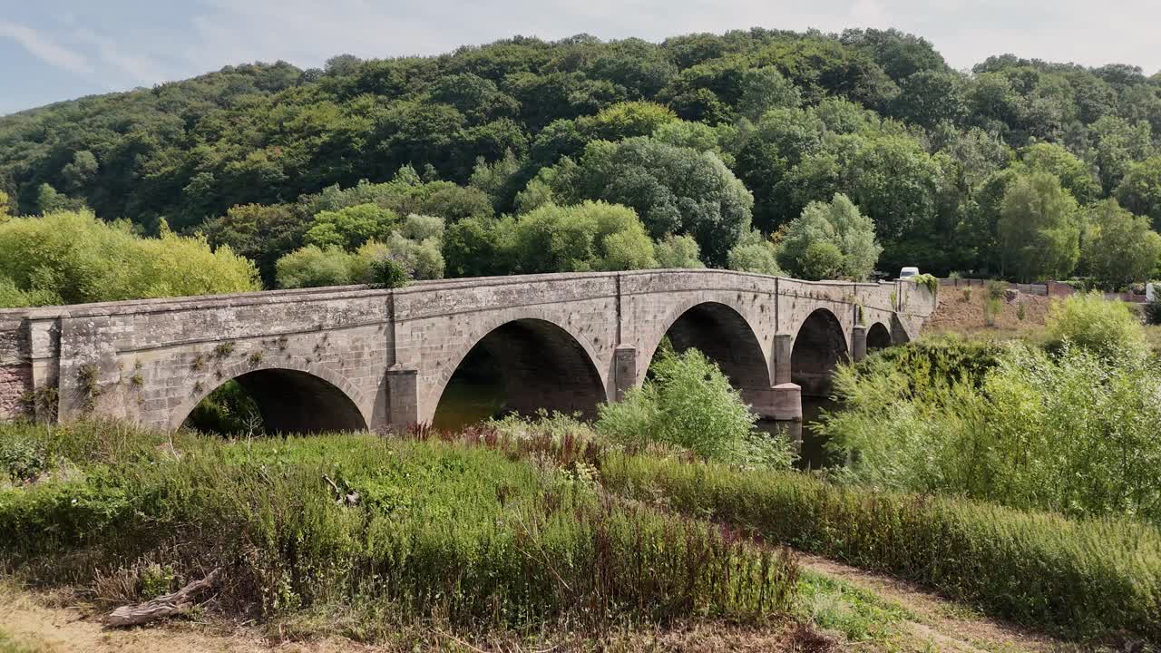 Fiume Wye, Valle di Wye, Herefordshire. Volo lento e basso verso un antico ponte di pietra sul fiume con acque calme, sotto uno splendido sole estivo e uno sfondo boscoso.