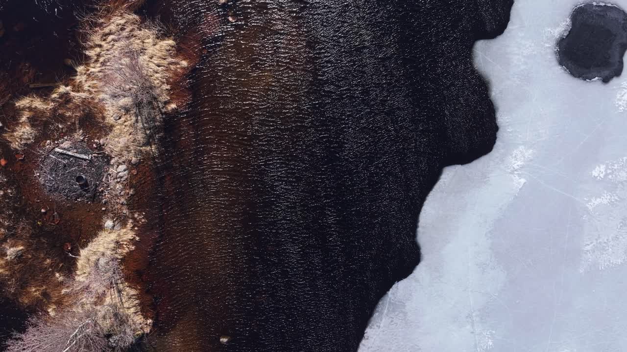 Rippled water around rocky shallow shoal. Lake partially covered in ice. Sunny Spring weather. Charred fire pit in center. Aerial top down view.