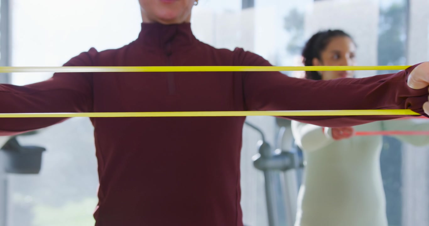 Due donne che si allenano in palestra, la donna in primo piano inizia ad afferrare le fasce, allungandole verso l'esterno per tonificare il petto