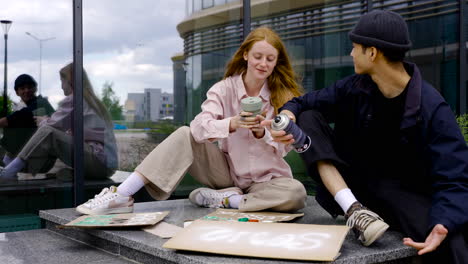 Ginger-Woman-Drinking-Coffee-While-Man-Painting-Board-For-Protest